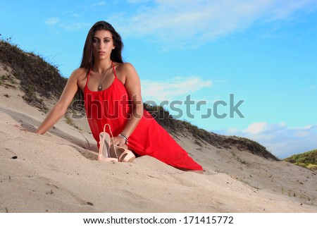 A beautiful brunette lady dresses in a long flowing red dress is resting on a dune on a beach in Africa.