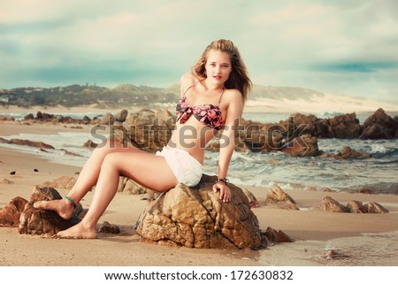 A beautiful blond is lying on her stomach on a rock on the beach in Africa. She has a bikini and white short pants on, the beach can be anywhere in South Africa or Mozambique or an island