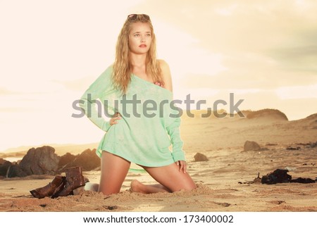 A beautiful blonde lady is sitting on her knees on the beach with the setting sun throwing golden colours behind her. she has her boots next to her.