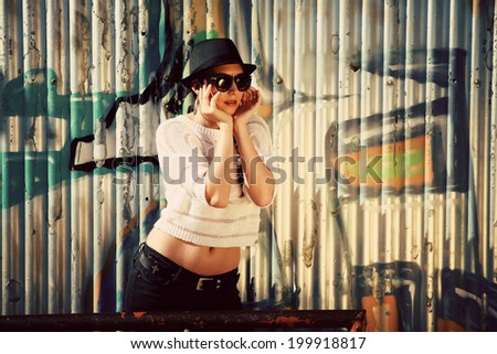 A beautiful young brunette lady is posing in front of a tin building with graffiti all over it.