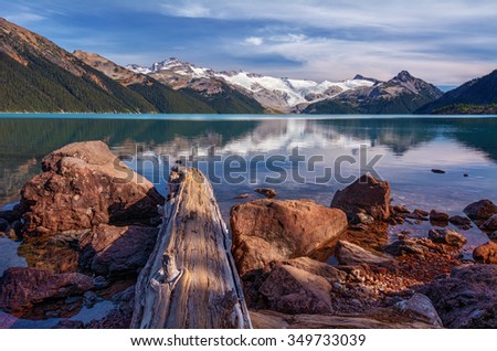Snow-capped mountains, calm glacial lake, rocks and log on the foreground