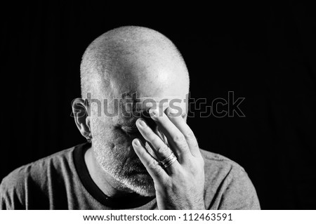 Black and white shot of a middle aged bald man with facial hair holding his head in his hands with a black background