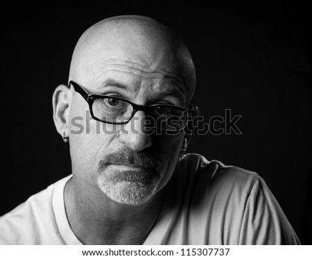 black and white head shot of a middle aged stern looking bald man with facial hair looking into camera wearing glasses with a black background