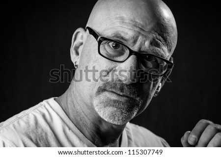 black and white head shot of a middle aged stern looking bald man with facial hair looking into camera wearing glasses with a black background