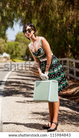 Young woman in a vintage dress with a vintage piece of luggage standing on the side of the road in the country waiting for someone