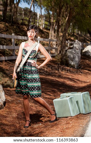 Full length color shot of a young woman in a vintage dress standing on a country roadside with a black and white electric guitar and vintage luggage