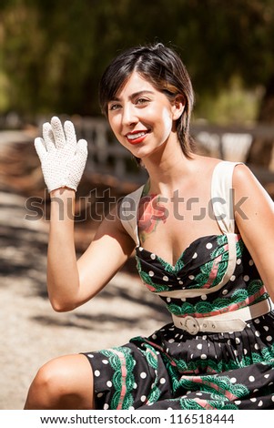Young girl on the side of the road sitting on vintage luggage wearing a vintage dress waving to someone while wearing white gloves