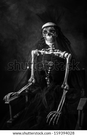 A black and white three quarter shot of a skeleton in a black wedding dress sitting in a directors chair with a necklace and a black veil.