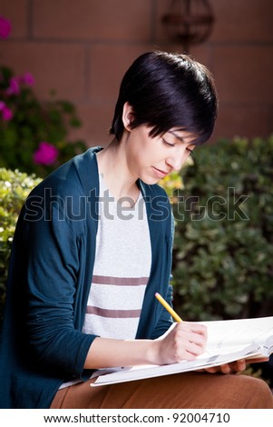 Young girl studying for test writing in a textbook sitting on the wall in the backyard
