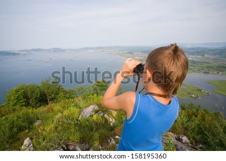 Boy looking through binoculars from the top of the mountain to the city by the sea.