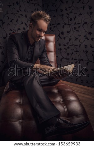 Handsome young man in dark suit relaxing on luxury sofa with book.