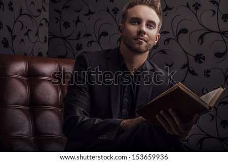 Handsome Young Man In Dark Suit With Book Relaxing On Luxury Sofa.