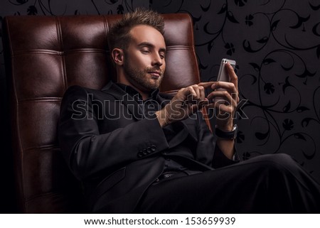 Handsome Young Man In Dark Suit Relaxing On Luxury Sofa With Mobile Phone.