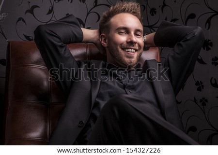 Handsome happy young man in dark suit relaxing on luxury sofa.