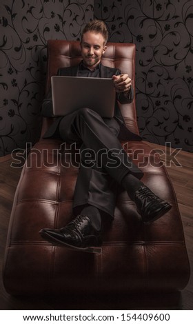 Handsome smiling young man in dark suit with note book relaxing on luxury sofa.