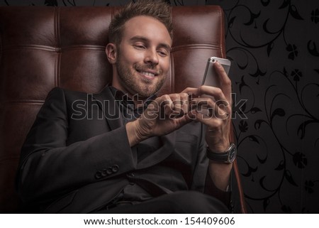 Handsome smiling young man in dark suit with phone relaxing on luxury sofa.