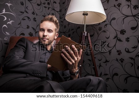 Handsome young man in dark suit with book relaxing on luxury sofa.
