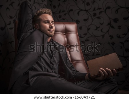 Handsome young serious man in dark suit with book relaxing on luxury sofa.