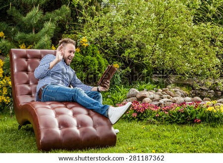Young handsome man in casual clothes sit in luxury sofa with iPad in summer garden.
