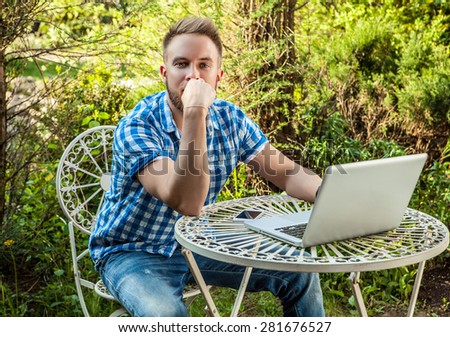 Young smiling handsome man in casual clothes work at an iron table with computer against country garden.