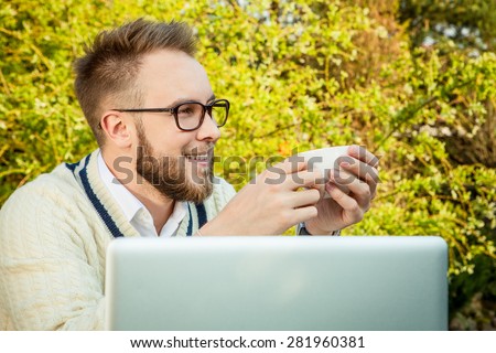 Young stylish handsome man in bright confidential clothes work at  iron table with computer & teapot against country garden.