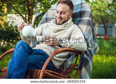 Handsome man relax in rocking-chair with teapot & cup in summer garden.