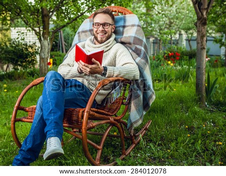 Handsome man relax in rocking-chair & reading red book in summer garden.