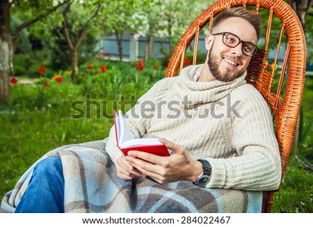 Handsome man relax in rocking-chair & reading red book in summer garden.