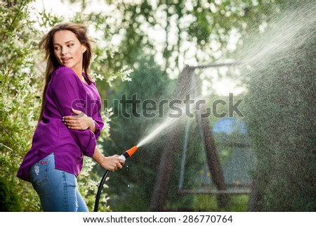 Beautiful joyful young girl in violet shirt poses in a summer garden with a water hose.