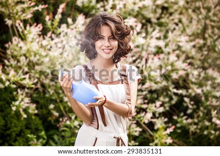 Outdoors portrait of beautiful & positive young woman in overalls which posing with water spray in solar summer garden.