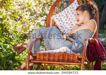 Outdoors portrait of beautiful young woman which sitting in rattan rocking-chair in evening summer garden.