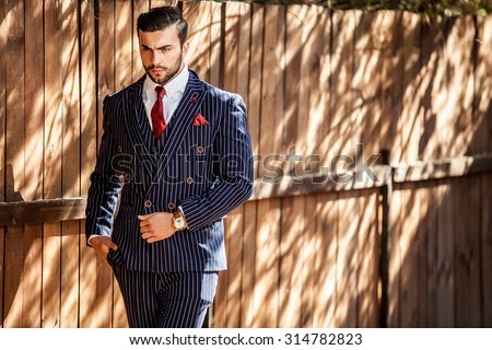 Elegant handsome man in classical blue suit poses near  wooden fence.