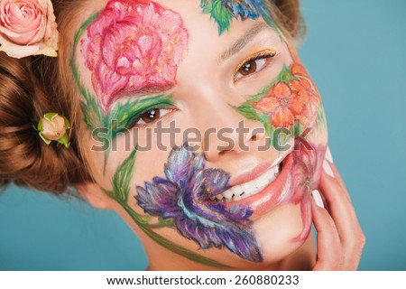Spring portrait of smiling female model with hand drawing flowers on her face. Color face art woman. On blue background