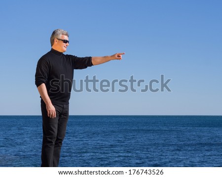 Handsome man standing in profile in a stylish black outfit pointing ahead of him at copy space while at the seaside with a calm blue ocean backdrop