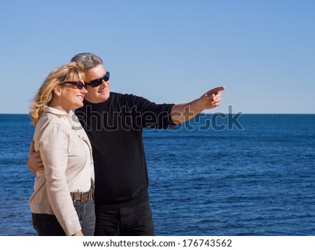 Affectionate mature couple at the seaside standing arm in arm with the husband pointing out something to his wife against a calm blue ocean on a hot sunny day