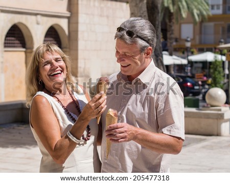 Laughing mature senior couple eating ice cream together while he try to steal her ice cone on a hot summer day as they stand in a tropical town square