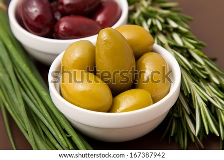 Bowls filled with pickled olives on clay plate. With lying side rosemary and chives.
