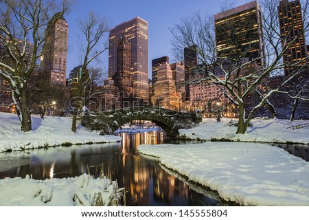 New York City Manhattan Central Park In Winter With Snow, Gapstow Bridge; Freezing Lake And Skyscrapers At Dusk.