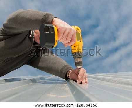 Man using  an electric drill to fasten down a metal roof, view from below, up against partly cloudy sky