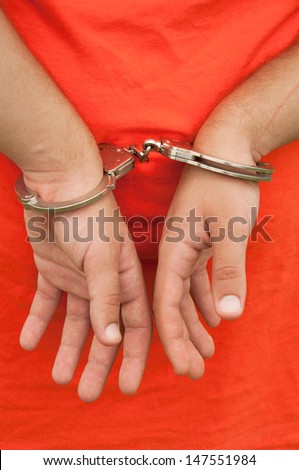 Close-up image of hands in handcuffs behind a person\'s back against an orange shirt or jumpsuit
