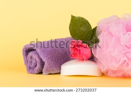 Soap topped with pink rose next to a shower puff and wash cloth against light yellow background