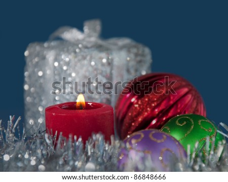 Christmas candle burning in the middle of silver tinsel, surrounded by colorful ornaments, against blue background