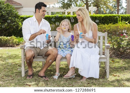A young family mother & father parents with girl child blowing bubbles having fun together sitting on a bench in a sunny park or garden.