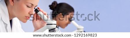 Panoramic web banner of female medical or scientific researcher or doctor using her microscope in a laboratory with her Asian colleague out of focus behind her.