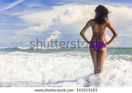 Rear view of a beautiful young woman in bikini standing in the surf waves on a deserted tropical beach with blue sky