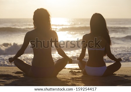 Two sexy young women or girls wearing bikinis sitting cross legged on a deserted tropical beach at sunset or sunrise