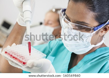 A female Asian medical or scientific researcher or doctor using a pipette and sample tray to test blood sample in a laboratory with her blond female colleague out of focus behind her.