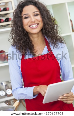 A beautiful happy young woman or girl wearing a red apron & using a tablet computer while cooking in her kitchen at home