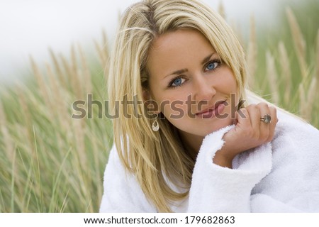 A beautiful blond haired blue eyed model wearing a white toweling robe sits amid tall grass
