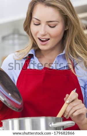 A beautiful blond girl or young woman looking happy wearing a red apron & cooking in her kitchen at home